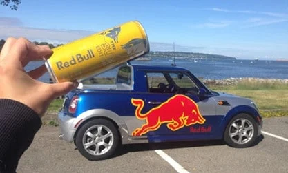Person, die Red Bull hält, kann über einem Red Bull-Miniauto stehen