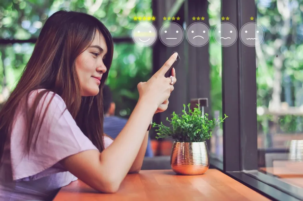 Akıllı telefonunda müşteri deneyimini derecelendiren bir kadının profil görünümü. Sağ üst köşedeki şeffaf gülen yüz grafikleri, mutludan üzgüne değişir.