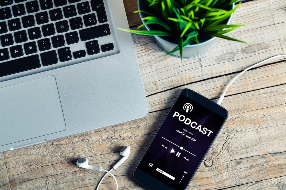 Podcast jugando en un teléfono móvil al lado de una computadora portátil abierta y una planta interior en un escritorio de madera.