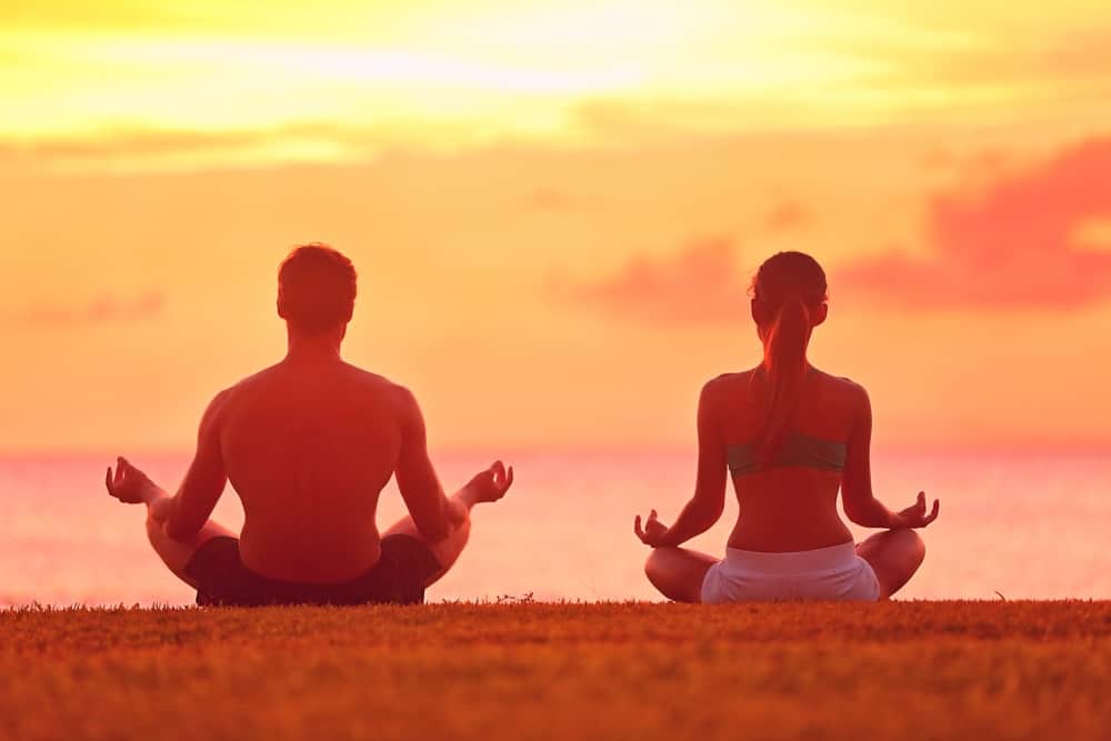 Vista posteriore di un uomo e di una donna che fanno una posa di meditazione a terra di fronte al cielo al tramonto.