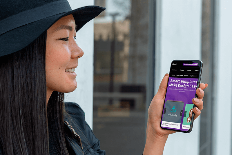 Modell eines lächelnden Mädchens mit einem Hut, der ihr Iphone Xs Max in einem urbanen Szenario hält