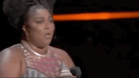 Gif celebritate. Lizzo poartă o rochie strălucitoare fără mâneci, dă din cap cu insistență și pompează în aer o statuetă de premiu în timp ce strigă: „Hai!