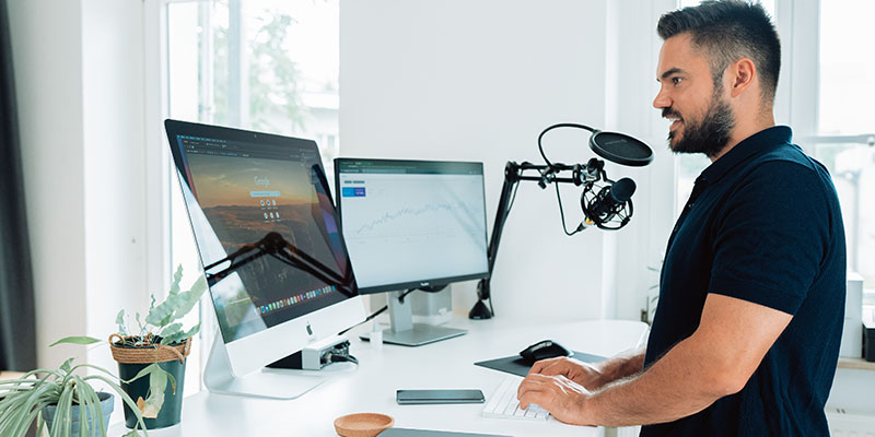 Homem trabalhando com microfone de podcast em frente a uma mesa e duas telas com computador de tela dividida e laptop na mesa de uma agência de mídia social.