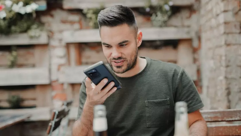 男性がテーブルに座りながら、携帯電話を見ながらスピーカーを使って話しています。